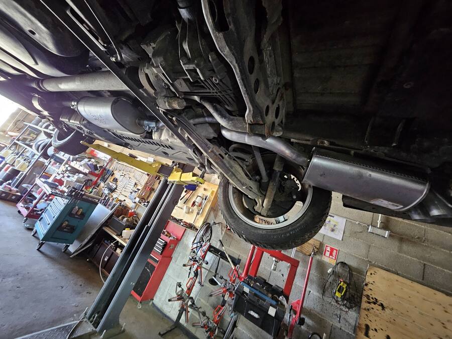 Dessous d'une voiture levée sur le pont élévateur de l'atelier Aurél auto, avec l'établi et les outils au fond